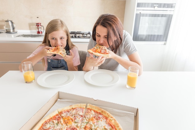 Mãe e filha sentadas na cozinha a comer pizza e a divertir-se concentradas na filha.