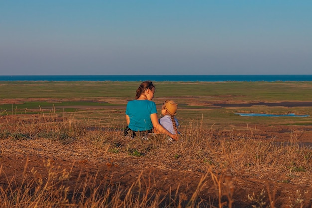 Mãe e filha sentadas perto da paisagem natural dos lagos Uma garotinha com uma longa trança de cabelo loiro abraçada com a mãe ao ar livre