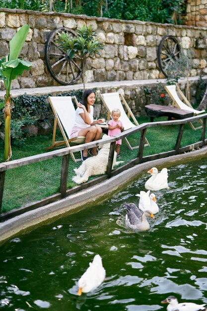 Mãe e filha sentam-se em cadeiras no gramado e alimentam os patos no lago