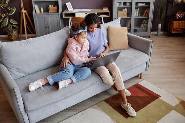 Mãe e filha usando laptop juntos sentados no sofá em casa aconchegante