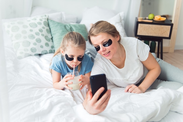 Mãe e filha, usando tapa-olhos, bebem smoothie e tomam selfie ou videochamada de spa e procedimentos de cuidados com o corpo