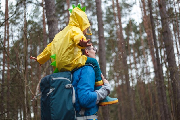 Mãe e filho andando na floresta depois da chuva em capas de chuva juntos a criança está sentada nos ombros da mãe vista traseira