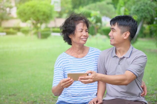 Mãe e filho asiáticos de meia-idade se olhando e olhando para um smartphone com um sorriso e sendo felizes no parque É um calor impressionante