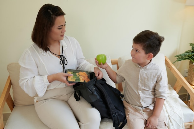 Mãe e filho colocando recipiente com merenda escolar e lanches na mochila escolar