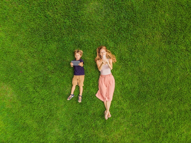 Mãe e filho estão deitados na grama do parque Mãe olha para o telefone filho olha para o tablet Fotos do drone quadracopter