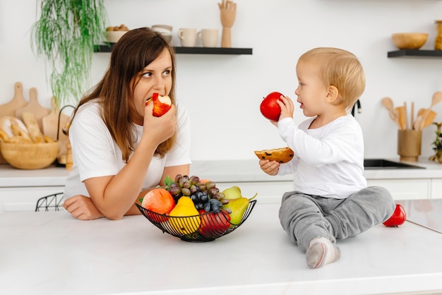 Mãe e filho estão na cozinha comendo comida saudável o bebê está segurando um biscoito e uma maçã vermelha