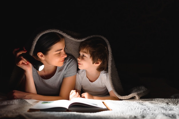Mãe e filho lendo um livro com uma lanterna debaixo do cobertor