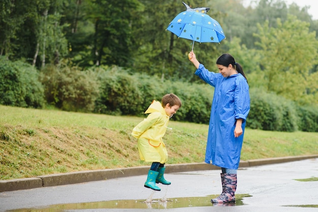 Mãe e filho, menino, brincando na chuva, usando botas e capa de chuva