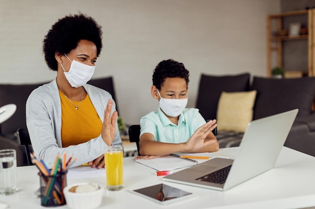 Mãe e filho negros com máscaras faciais acenando enquanto seguem aulas on-line em casa