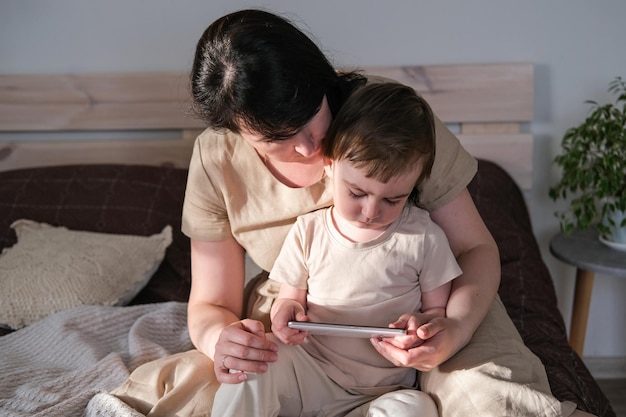 Mãe e filho pequeno sentam na cama e assistem desenhos em um smartphone