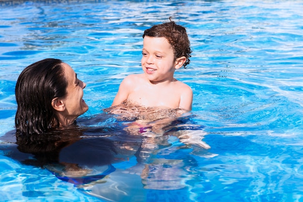 Mãe e filho tomando banho na piscina - ambos sorrindo alegremente, olhando um para o outro enquanto desfrutam