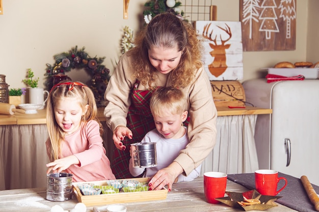 Mãe e filhos estão preparando cupcakes na cozinha. Decorações de Natal, tradições familiares, comida de Natal, vésperas de férias.