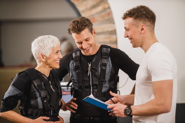 Mãe e seu filho adulto estão conversando com o personal trainer antes do treino EMS na academia.