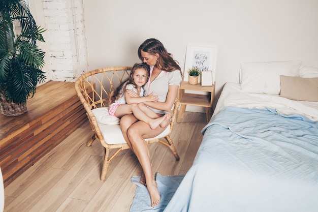 Mãe e sua filha pré-escolar linda filha de pijama, abraçando-se sentado em uma cadeira de vime pela manhã.