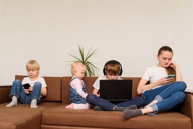 Mãe e três filhos sentam no sofá e usando telefone e laptop. Dependência de pessoas e tecnologia