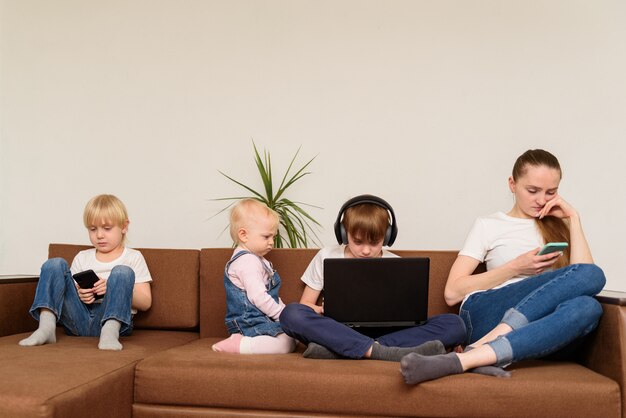 Mãe e três filhos sentam no sofá e usando telefone e laptop. dependência de pessoas e tecnologia