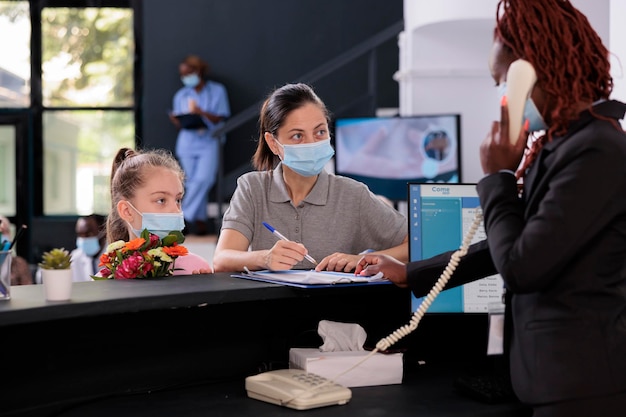 Mãe em pé no balcão com criança assinando papéis com seguro médico durante a consulta no saguão de espera do hospital. Pessoas usando máscara facial protetora para prevenir infecção por coronavírus