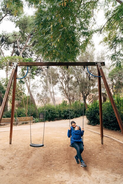 Mãe em um suéter azul quente e jeans balançando em um balanço com um bebê minúsculo em seu peito no parque