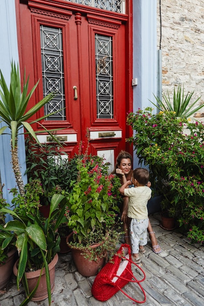Mãe em um vestido de verão colorido senta-se junto a uma porta vermelha envolvendo-se com seu filho pequeno em meio à flora exuberante de uma aldeia mediterrânea
