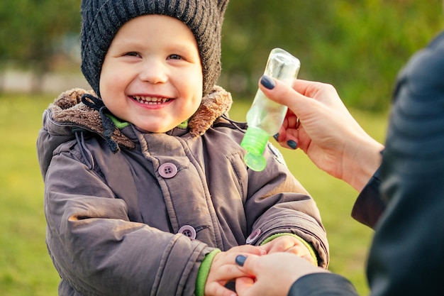 Mãe em uma jaqueta quente e chapéu de malha aplica um gel antisséptico gel antibacteriano menino andando no parque outono mulher loira elegante com uma criança contra o pano de fundo do lago