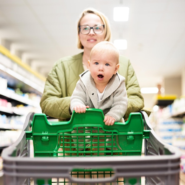 Mãe empurrando carrinho de compras com seu bebê bebê menino no corredor do departamento na mercearia do supermercado Comprar com o conceito de crianças