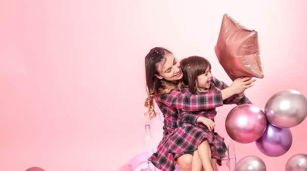 Mãe engraçada e criança sentada em uma parede transparente rosa cadeiras elegantes. Menina e mãe se divertindo com balões e confetes