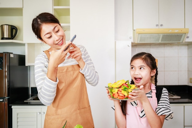 Mãe engraçada e garotinha gostam de tirar uma foto de salada de legumes atividade de lazer em uma cozinha