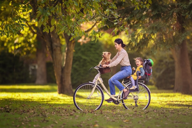 Mãe está andando de bicicleta no parque com seu bebê na parte de trás e seu cachorro na frente no pôr do sol