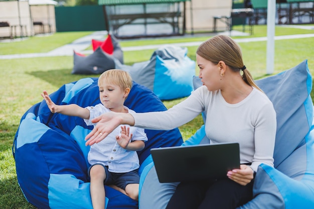 Mãe está em licença de maternidade Encantadora jovem com filho trabalhando ao ar livre no parque de verão com laptop enquanto está sentado na cadeira na grama verde