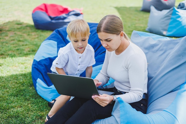 Mãe está em licença de maternidade Encantadora jovem com filho trabalhando ao ar livre no parque de verão com laptop enquanto está sentado na cadeira na grama verde