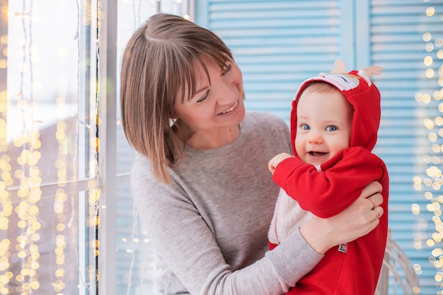 Mãe feliz brincando com seu filho em uma fantasia de veado de Papai Noel vermelho no fundo da janela e luzes guirlandas