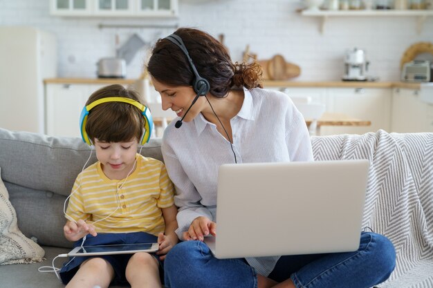 Mãe feliz com a criança sentada no sofá em casa durante o trabalho de bloqueio no laptop, criança brincando no tablet