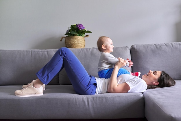 Mãe feliz com o filho da criança em casa. Mulher rindo brincando com o bebê deitado no sofá. Família, infância, amor, conceito de felicidade na maternidade