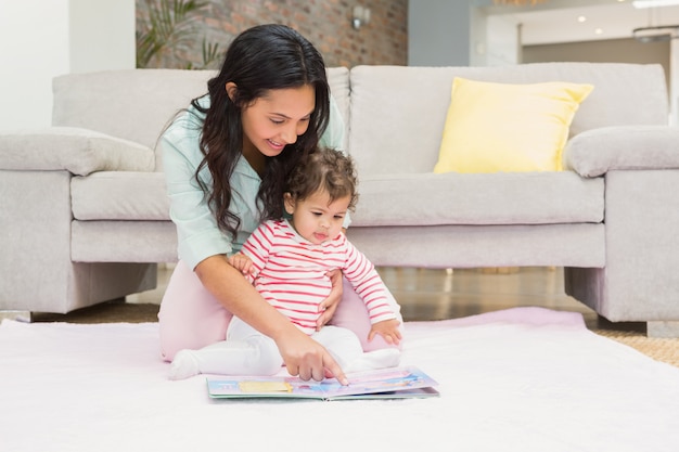 Mãe feliz com seu bebê olhando para um livro no tapete na sala de estar