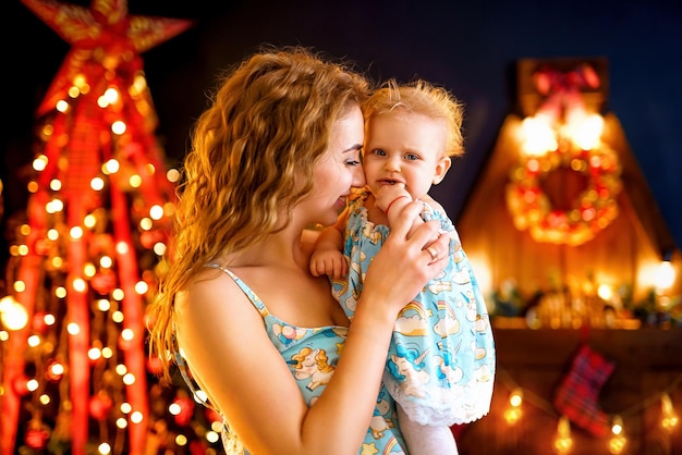 Mãe feliz com seu bebê parado perto da árvore de Natal na sala de estar com o mesmo pijama de Ano Novo