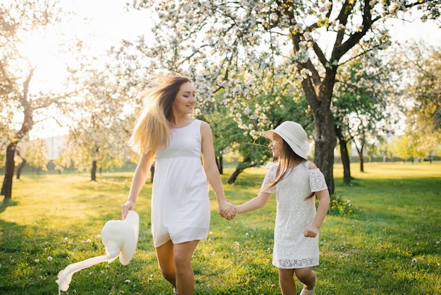 Mãe feliz e sua filha em vestidos brancos correm pelo jardim da primavera ao pôr do sol