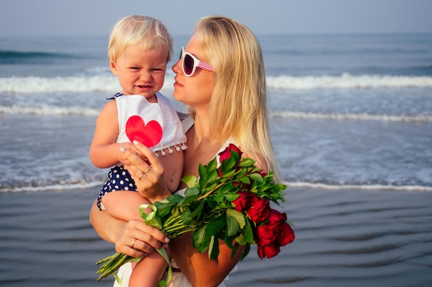 Mãe feliz em um vestido branco com a filha segurando um cartão postal de coração de papel e um buquê de flores rosas vermelhas na praia. Linda mulher loira e bebê mãe e dia dos namorados à beira-mar