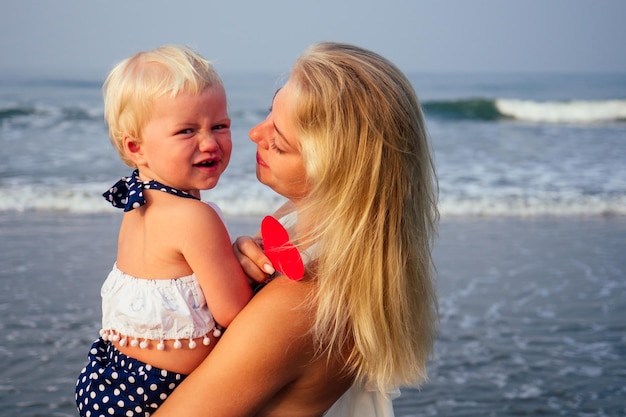 Mãe feliz em um vestido branco com a filha segurando um cartão postal de coração de papel na praia. Linda mulher loira e seu bebê comemorando o dia da mãe e dos namorados à beira-mar