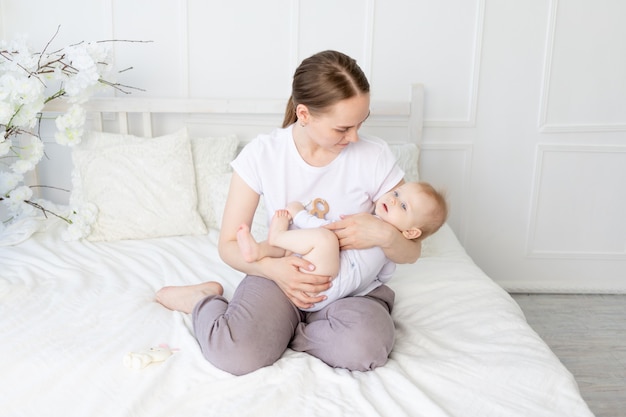 Mãe feliz segura o bebê nos braços com um roedor na boca em uma cama branca com lençóis de algodão em casa