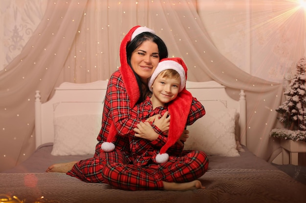 mãe filho com pijama de natal e chapéus de papai noel abraçando sentado na cama