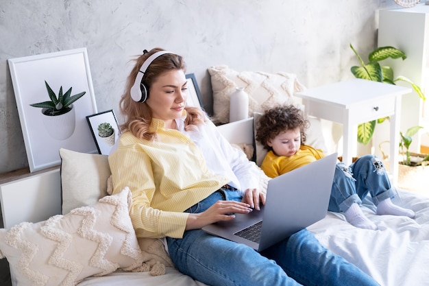 Mãe freelancer usando laptop em fones de ouvido sem fio deitada na cama com filha criança usando telefone