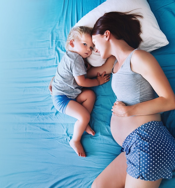 Mãe grávida e filho deitado na cama em casa. Grávida pela segunda vez com o primeiro filho. Menino e mãe da criança. Família na expectativa de um bebê. Gravidez, família, amor, conceitos de maternidade.