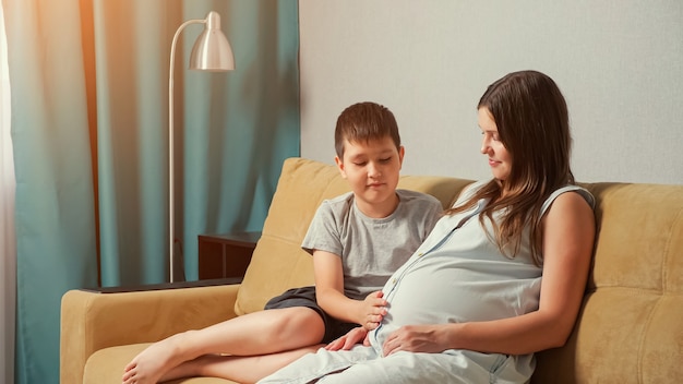 Mãe grávida e filho no sofá. Menino acariciando e abraçando a barriga.