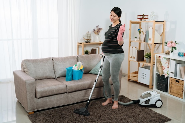 mãe grávida japonesa alegre de corpo inteiro está dançando música enquanto limpa o chão usando um aspirador em uma moderna sala de estar com luz do dia em casa.