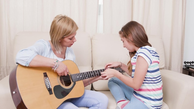 Mãe gutarista sentada no sofá com a filha ensinando quem deve tocar violão, apreciando a atividade musical. criança tocando instrumento musical durante a aprendizagem da lição de canto em casa. lazer educativo