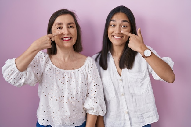 Mãe hispânica e filha juntas apontando com a mão dedo a cara e nariz sorrindo alegre conceito de beleza