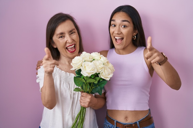 Mãe hispânica e filha segurando o buquê de flores brancas, apontando os dedos para a câmera com cara feliz e engraçada. boas energias e vibrações.