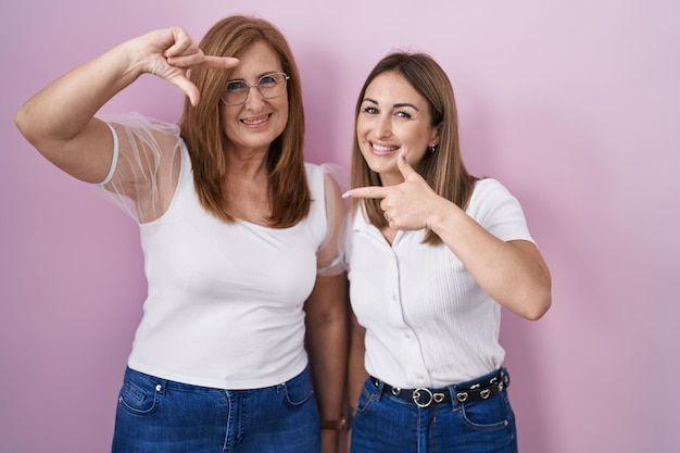 Mãe hispânica e filha vestindo camiseta branca casual sobre fundo rosa sorrindo fazendo moldura com mãos e dedos com criatividade de rosto feliz e conceito de fotografia