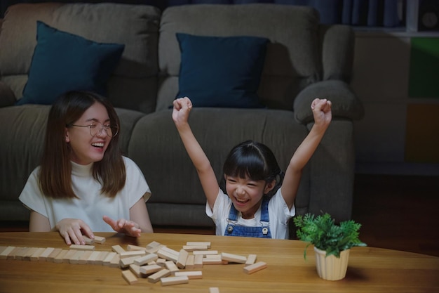 Mãe jovem asiática jogando jogo em bloco de madeira com sua filhinha na sala de estar em casa durante a noite, mulher sorridente ajuda a ensinar brincadeiras de criança construir construtor de blocos de madeira, educação