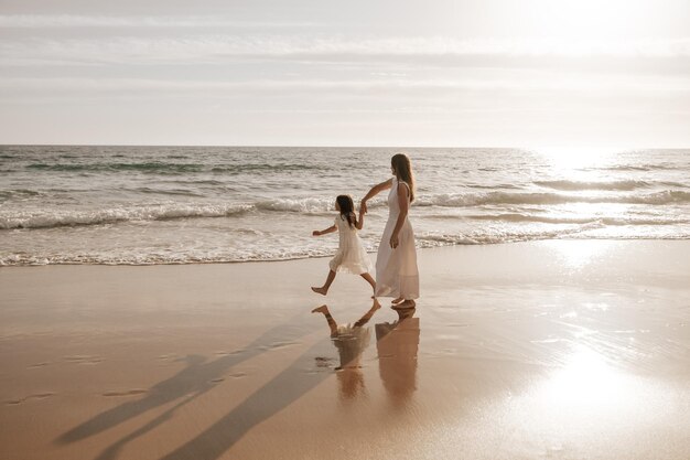 Mãe jovem com filha adorável em roupas semelhantes caminhando na praia de areia